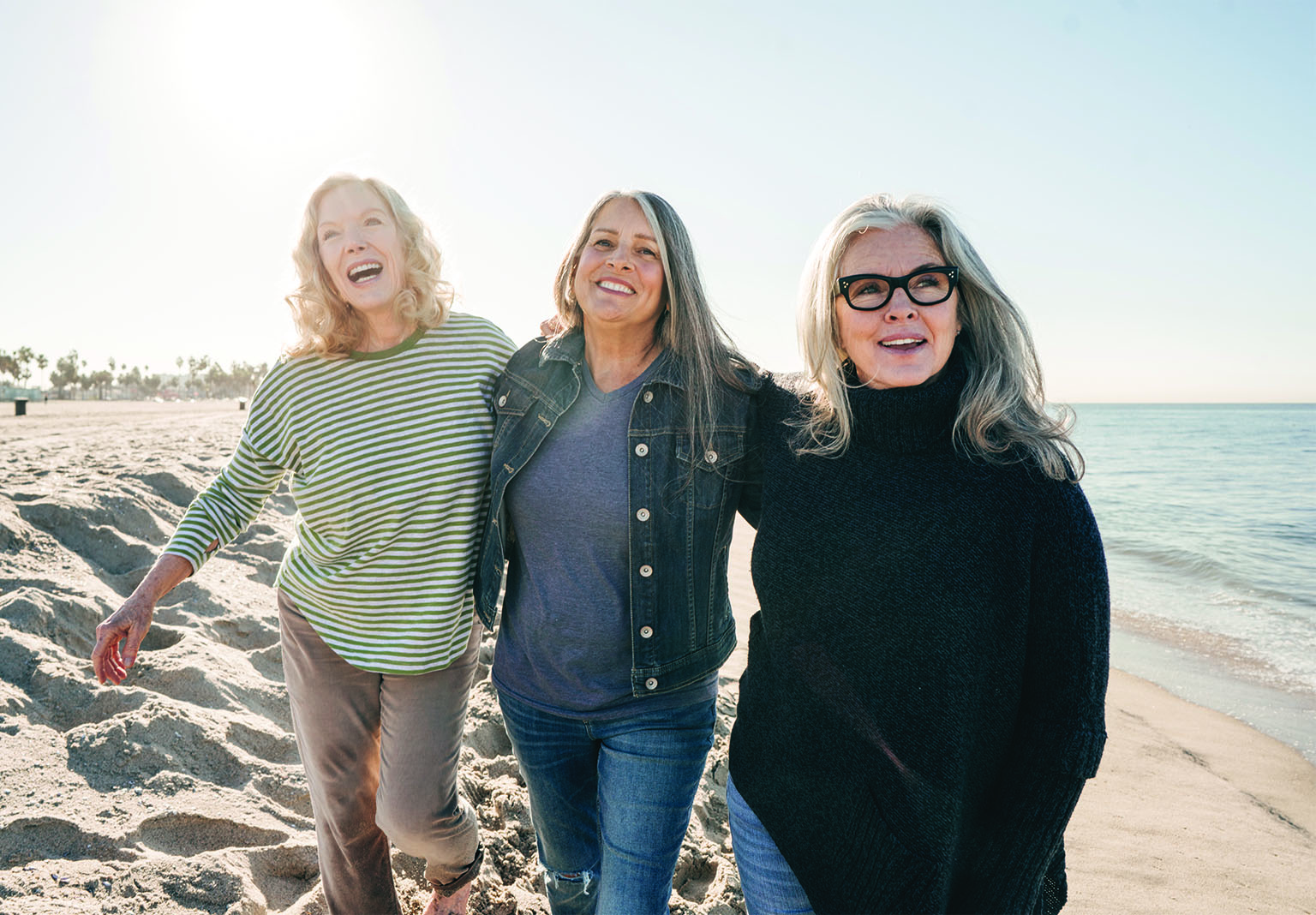 Three women walking arm-in-arm on a sunny beach, smiling and enjoying the day. They are dressed casually, with one in a striped shirt, one in a denim jacket, and one in a black turtleneck sweater. The ocean and clear sky provide a serene background.